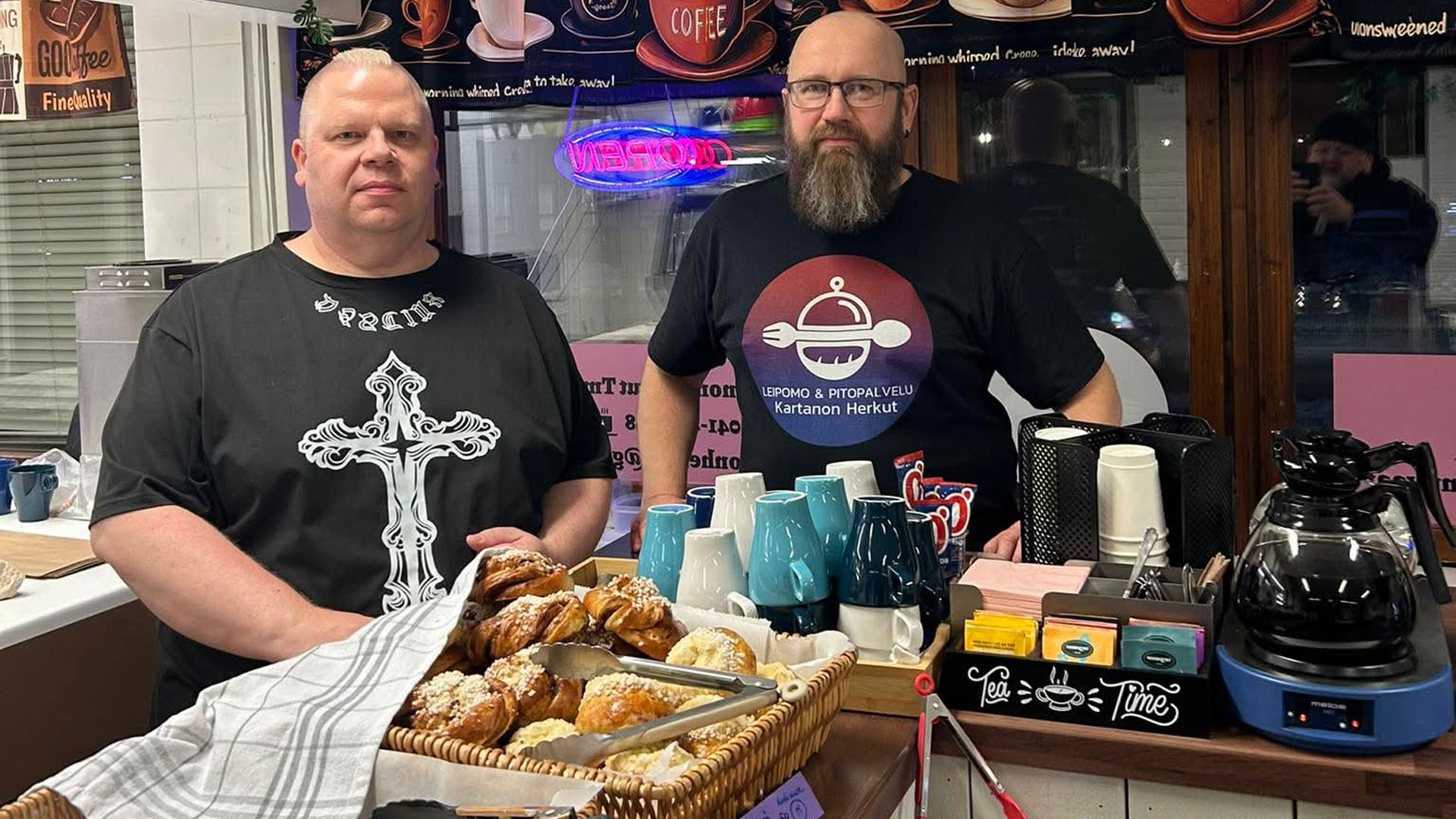 Kaksi miestä seisoo kahvilan tiskin takana, etualalla leivonnaisia ja kahvitarvikkeita.