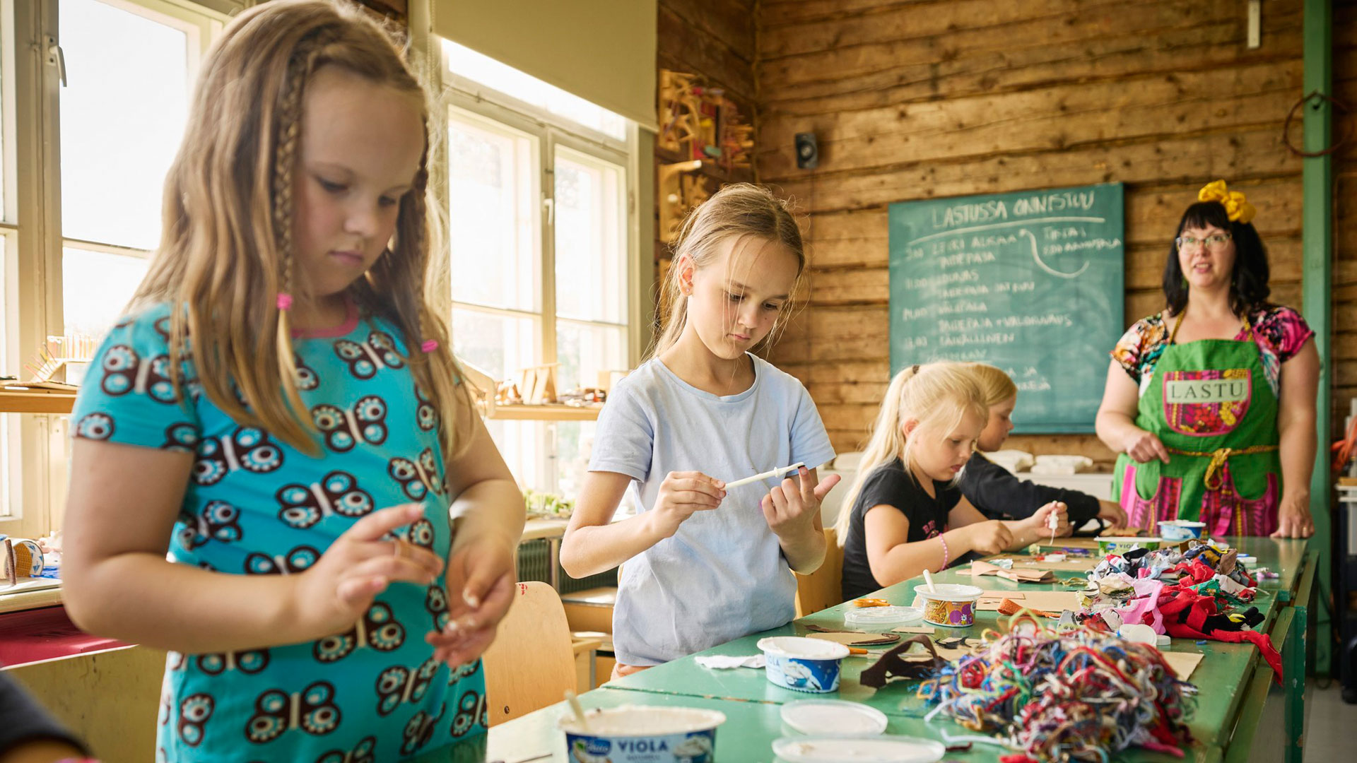 Neljä tyttöä askartelee. Taustalla nainen seuraa tekemistä.