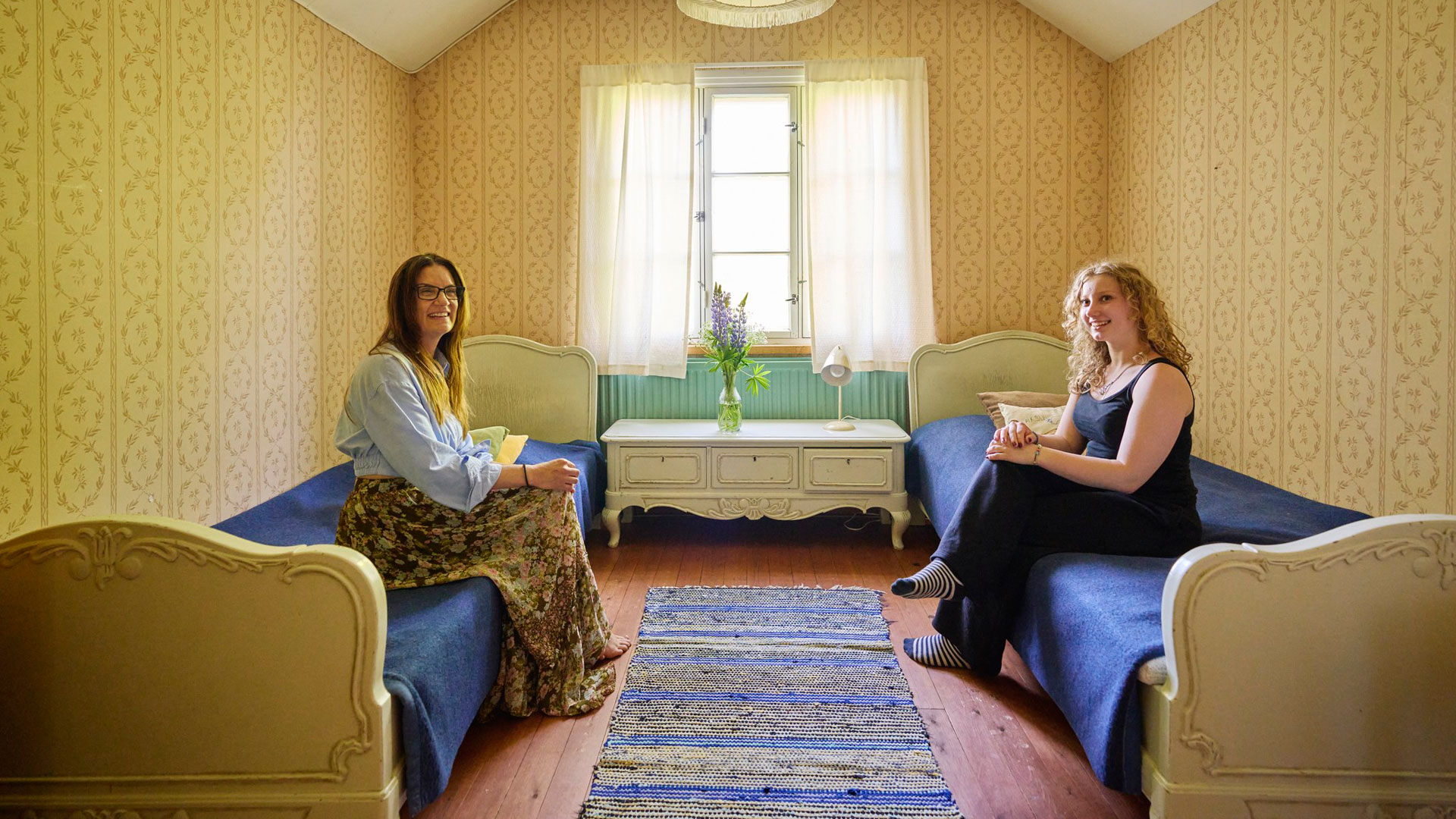 Kaksi naista istuu sängyillä huoneessa, jossa on lipasto, kaksi sänkyä ja räsymatto lattialla.