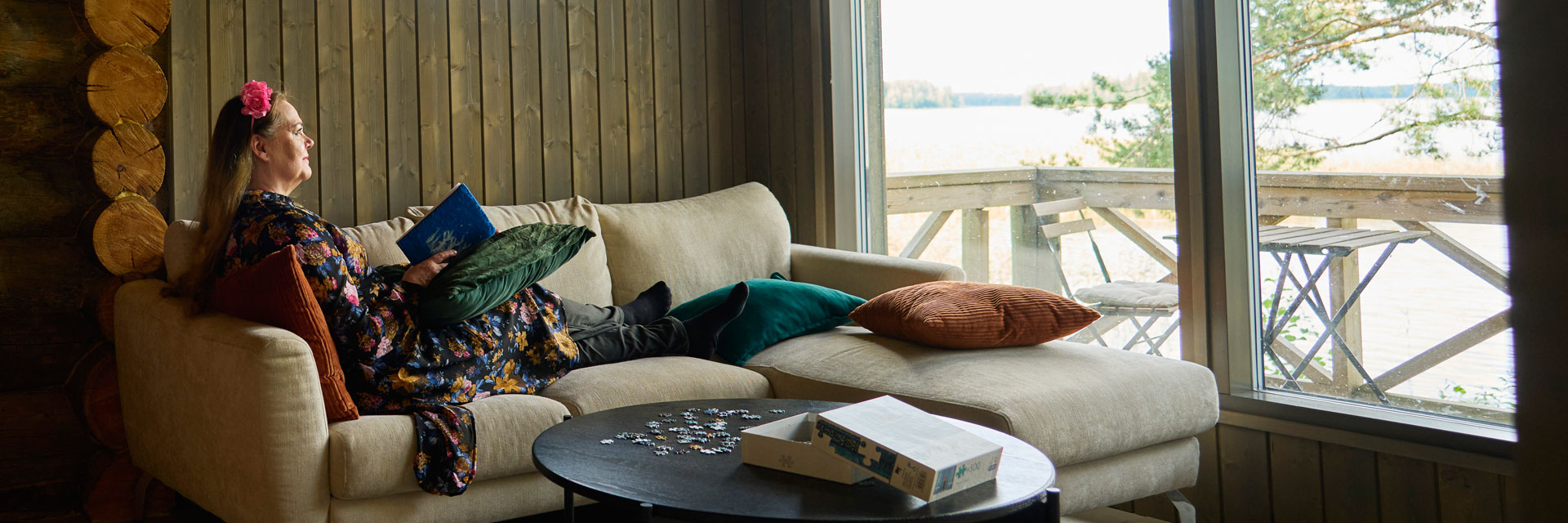 Nainen on mökin sisällä sohvalla kirjan kanssa. Ikkunan takana on järvimaisema.