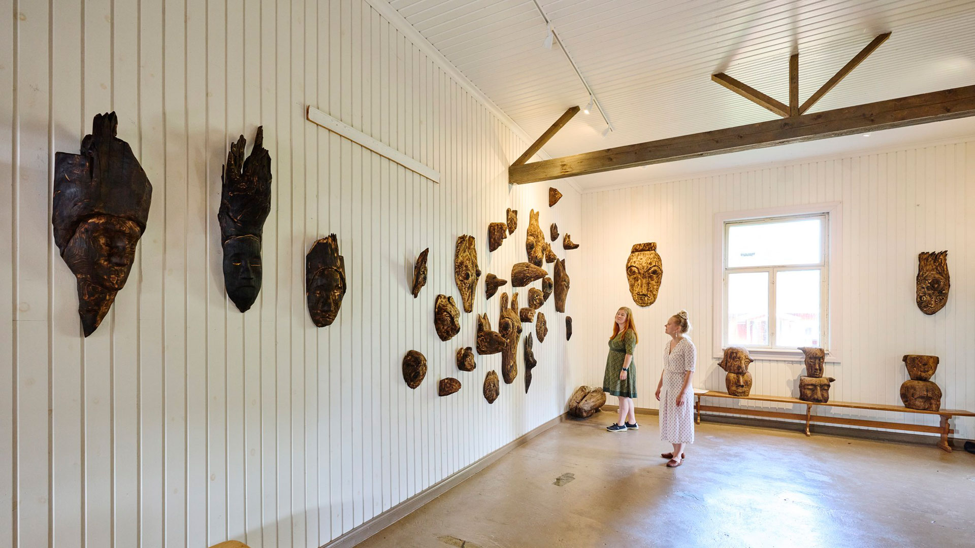 Kaksi naista katsoo seinälle ripustettuja puuveistoksia.
