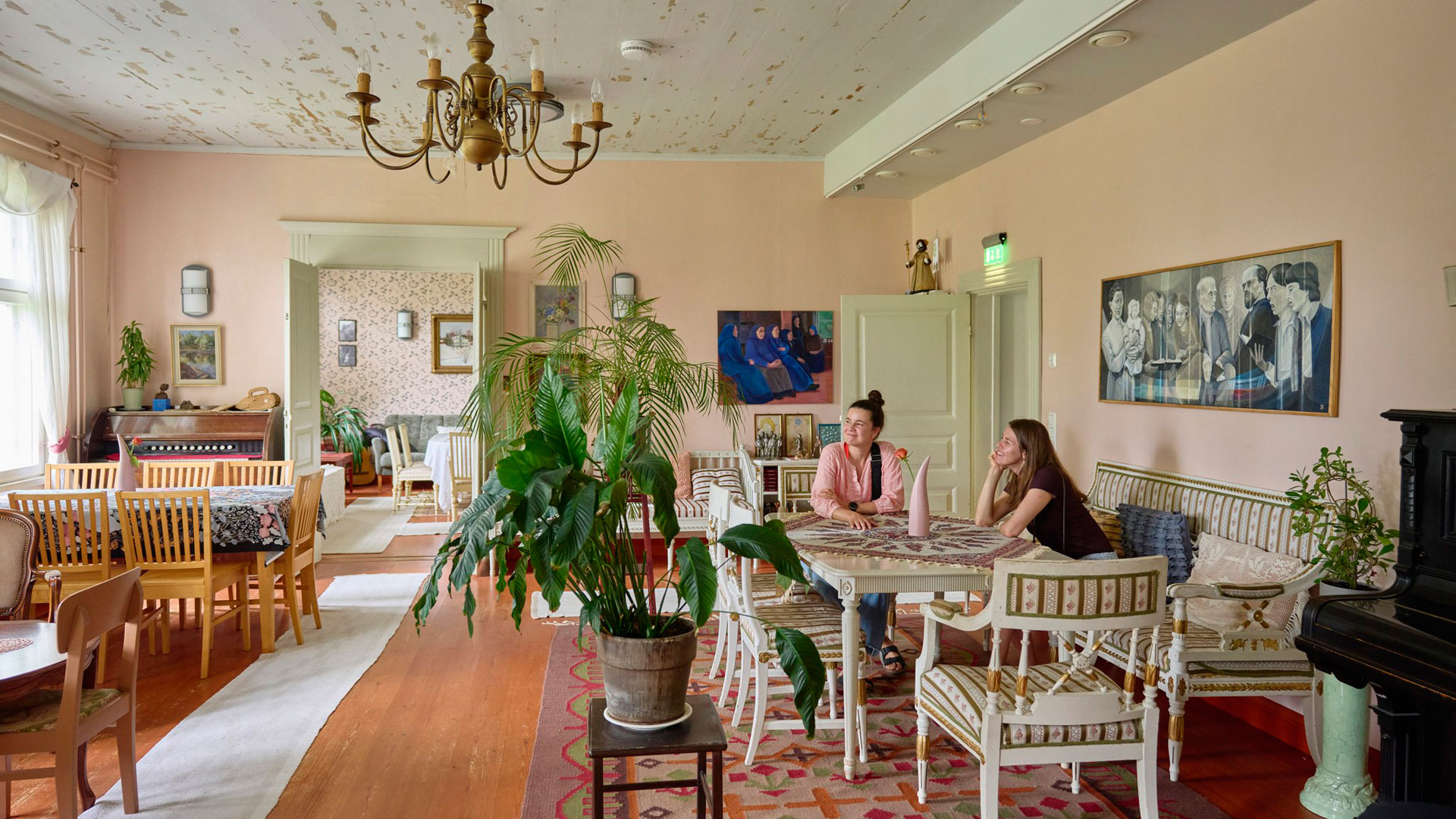 Kaksi hymyilevää naista istuu huoneessa pöydän ääressä ja katsoo ikkunaan päin.